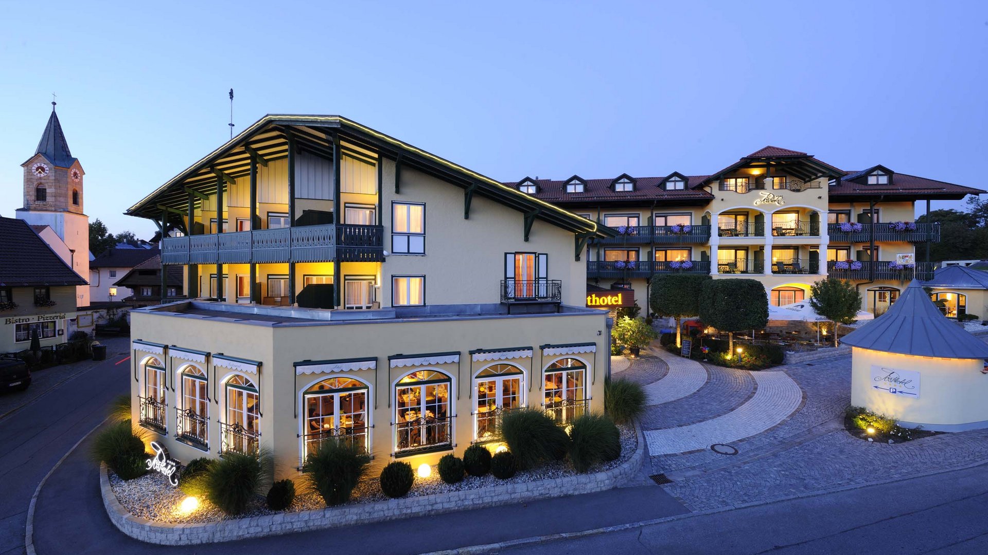 Wellness-Wochenende im Bayerischen Wald | Posthotel Rattenberg Beleuchtetes Hotelgebäude mit Terrasse und Dorfkirche bei Abenddämmerung