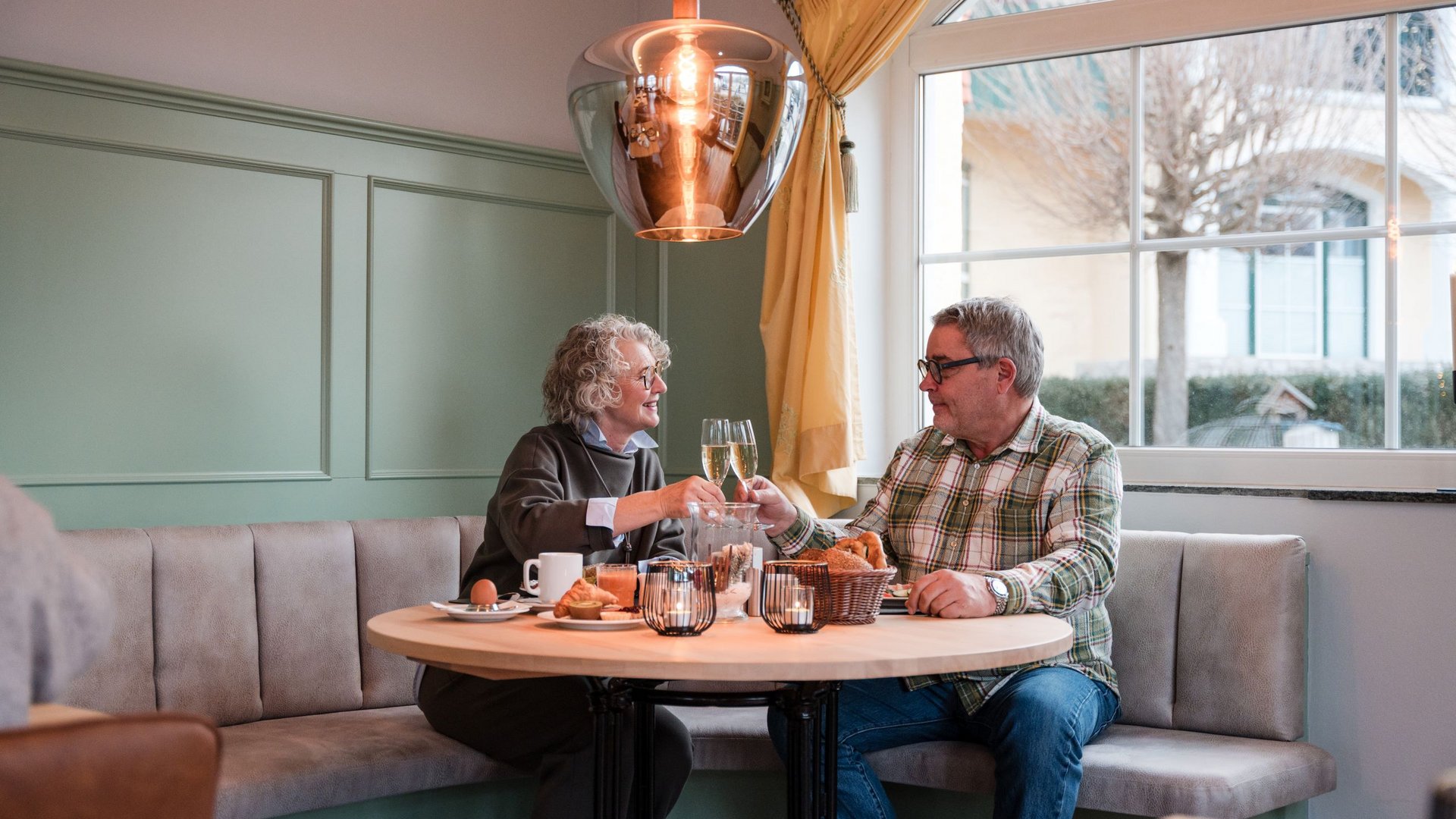 Romantisches Hotel in Bayern für pure Waldglücksmomente Älteres Paar stößt mit Sektgläsern bei Frühstück in gemütlichem Café an