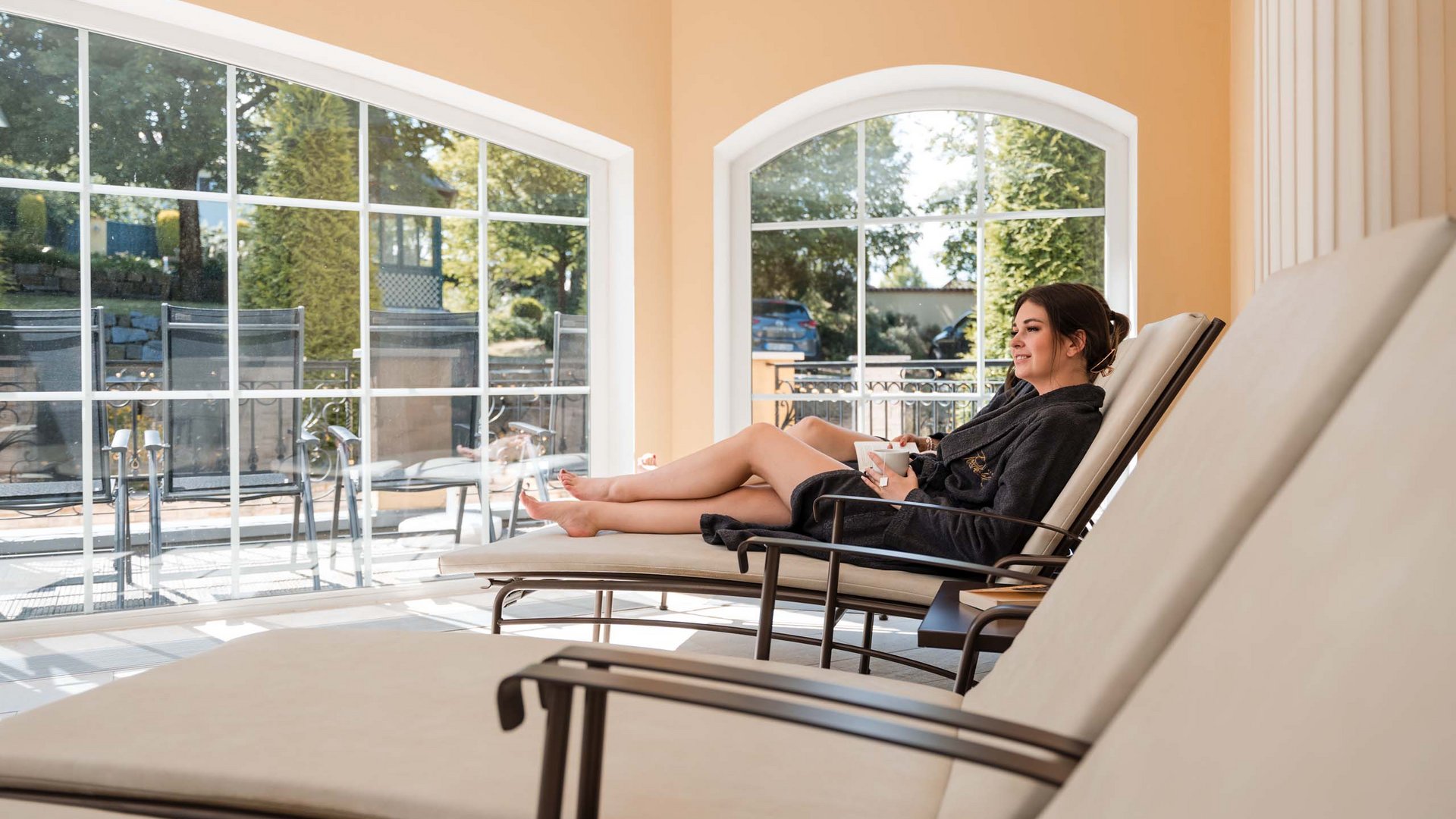 Day Spa: Machen Sie Tageswellness im Bayerischen Wald! Frau entspannt auf Liege mit Tasse in hellem Raum mit großen Fenstern