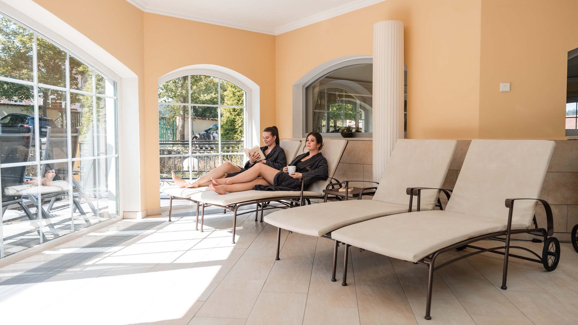 Ihr Hotel mit Hallenbad im Bayerischen Wald Zwei Frauen in Bademänteln entspannen auf Liegestühlen neben großen Fenstern