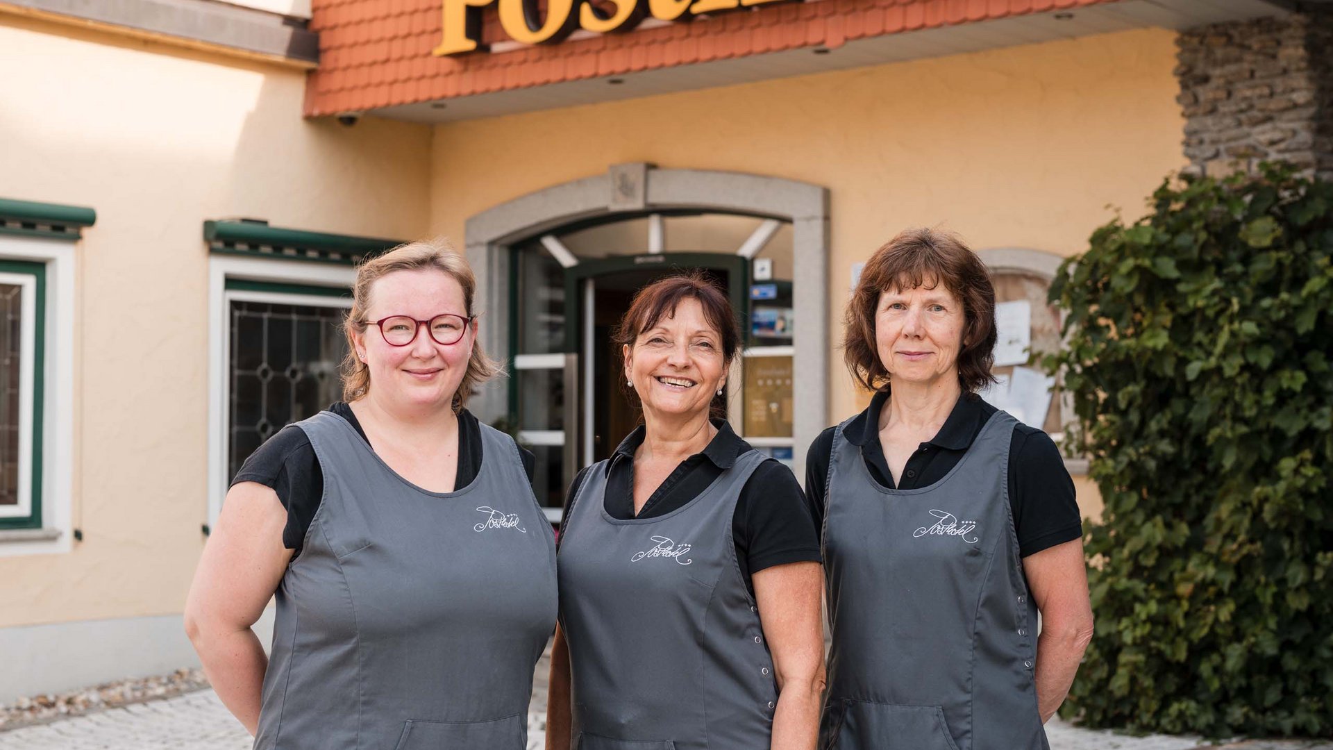 Ihr Arbeitsplatz mit Herz und Sinn Drei Frauen in grauen Schürzen vor einem Gebäude mit Posthotel-Schild